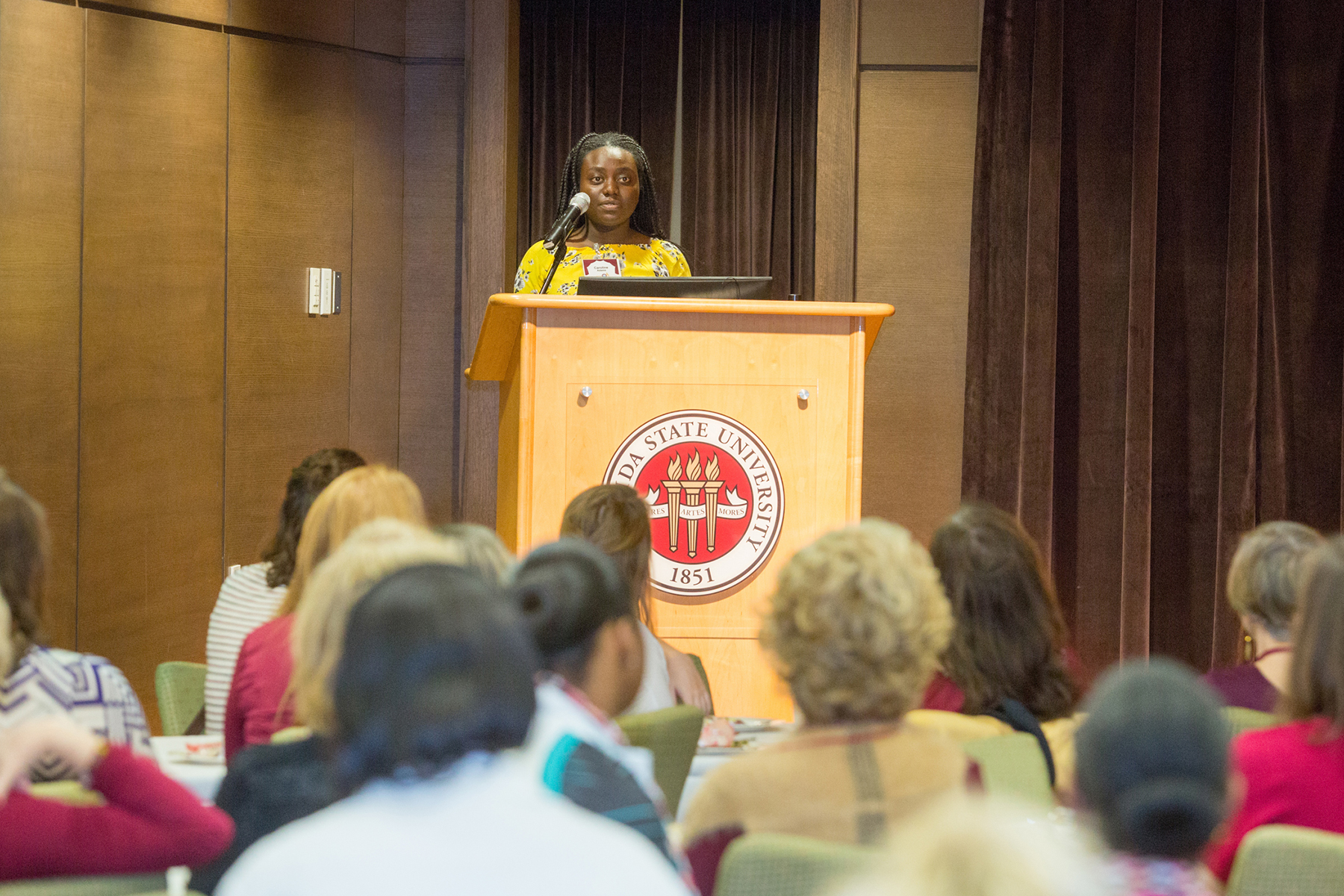 A photo of a young woman at a podium, speaking to an audience